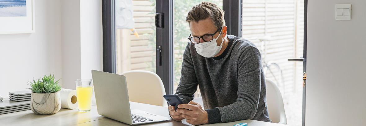Man wearing mask looking at cell phone and laptop