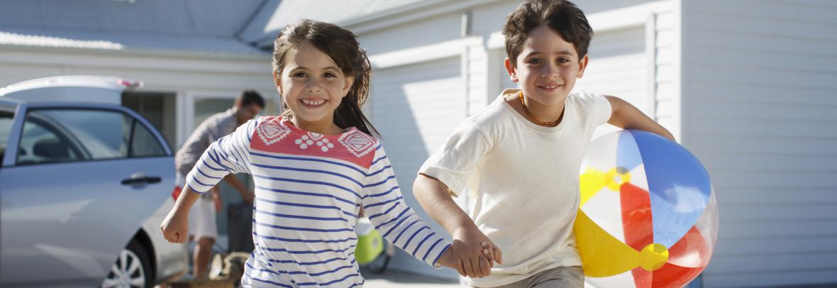Brother and sister kids with beach ball running toward beach with house in background