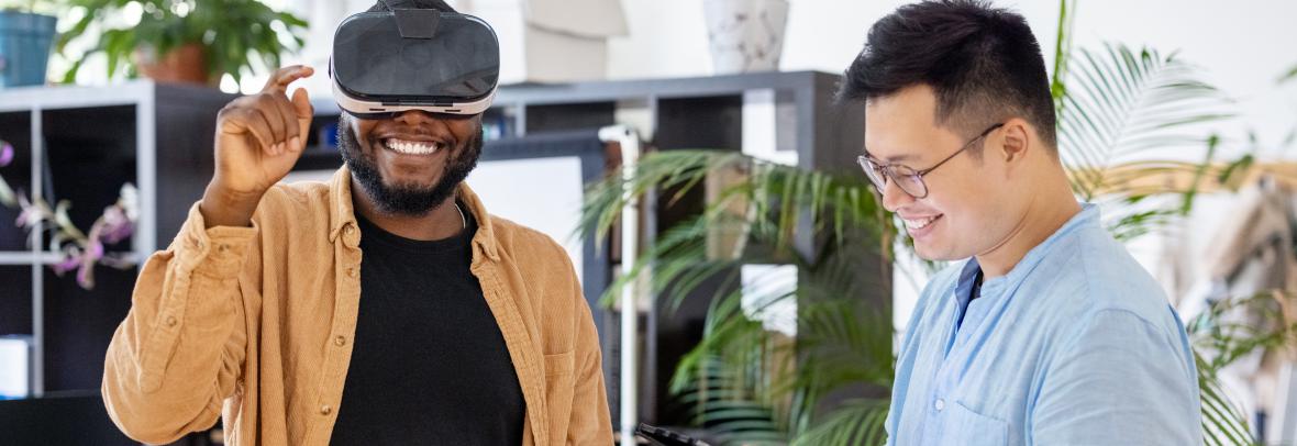 Colleagues testing out virtual reality device inside the metaverse - stock photo African man wearing virtual reality glasses and gesturing with ale colleague standing by using digital tablet in office. Men testing out virtual reality device at office.