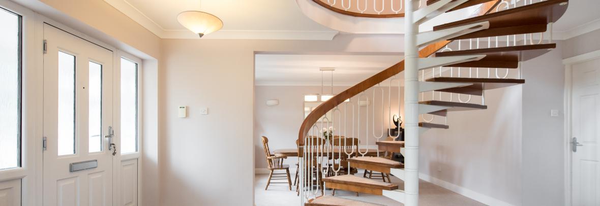 A general interior view of a retro style spiral staircase made of wood and metal in a open-plan hallway dining room with grey walls and beige carpet