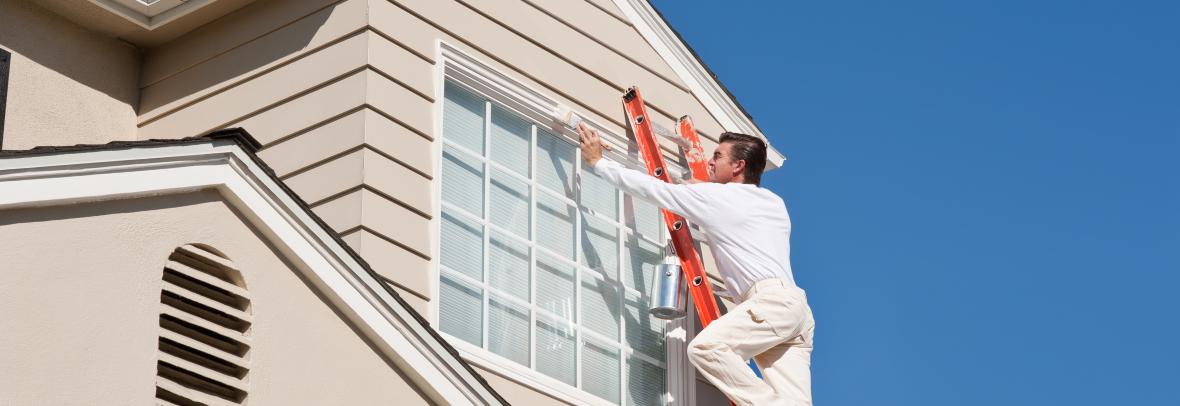 House painter with paint brush painting the trim of a 2nd story window of a Cape Cod Style house.