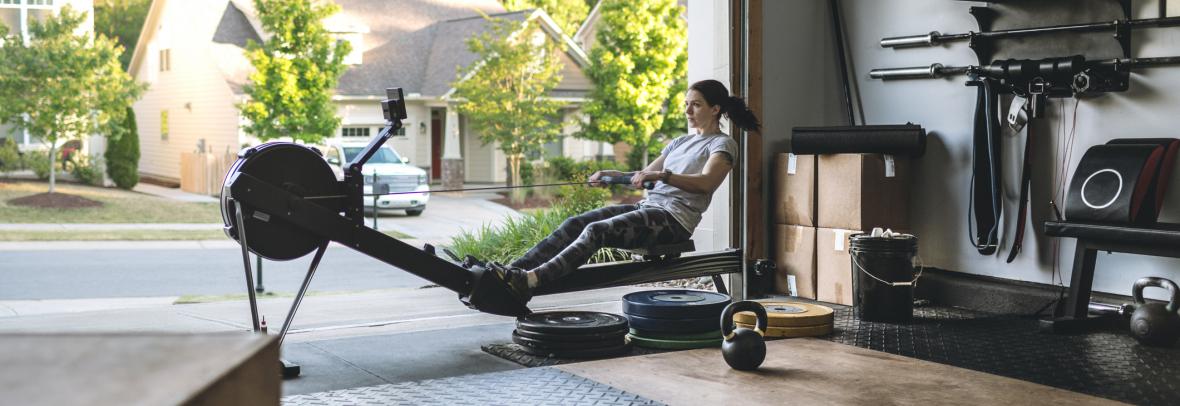 Photo of an active woman is exercising on a rowing machine in the home garage gym during covid-19 pandemic. Woman performing powerful pulls and focusing on endurance and cardio improvement.