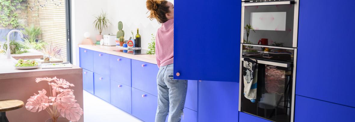 Full length view of woman in jeans and casual top standing on tiptoe to access high shelf in vibrant blue kitchen.
