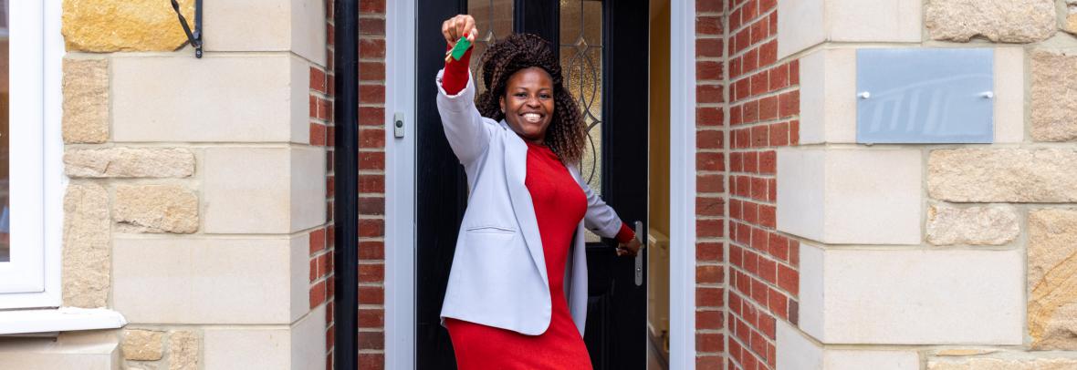 Three-quarter-length shot of a mature adult woman standing outside a vacant property. She is smiling happily at the camera, holding up a set of house keys in one hand whilst opening the door with the other. She is wearing smart casual clothing.