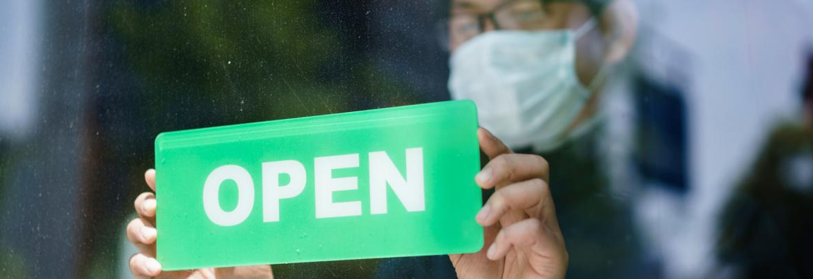store owner holding wearing mask holding open sign up to window