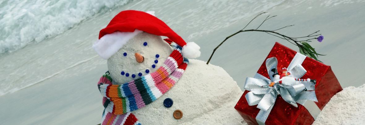 Snowman made of sand on the beach has a scarf and hat and Christmas gifts