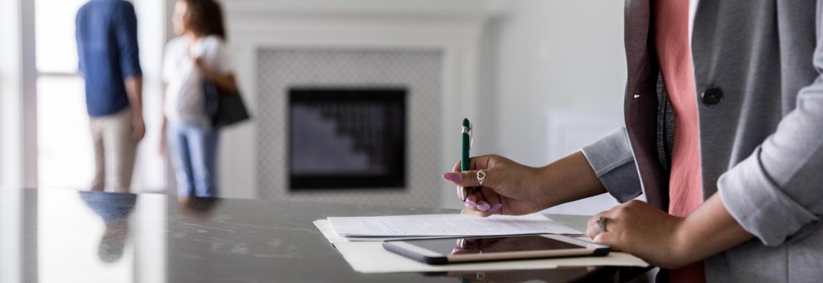Female real estate agent finishes up paperwork as a couple discuss the purchase of a new home in the background.