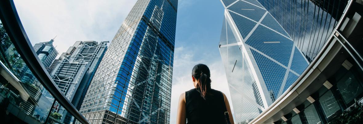 Rear view of professional young businesswoman standing against contemporary financial skyscrapers in downtown financial district and looking up into sky with positive emotion