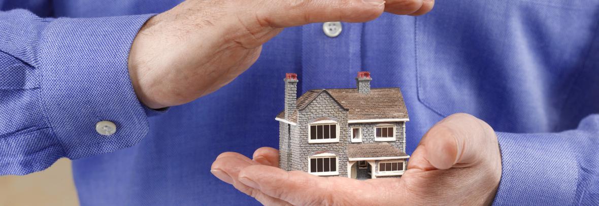 Model home in a man's one hand with his other hand covering it to suggest insurance