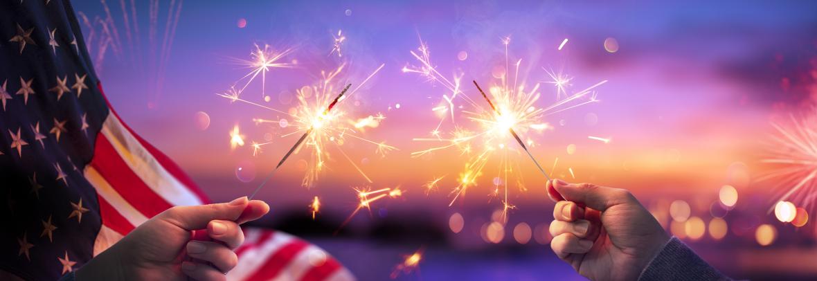 July 4 celebration hands holding sparklers with flag behind on left