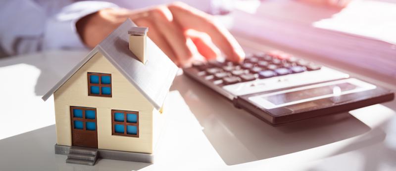 Photo of a persons hand using a calculator and a small house in the foreground