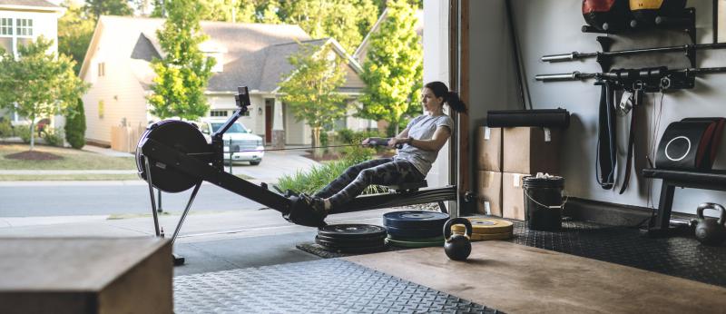Photo of an active woman is exercising on a rowing machine in the home garage gym during covid-19 pandemic. Woman performing powerful pulls and focusing on endurance and cardio improvement.