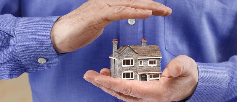 Model home in a man's one hand with his other hand covering it to suggest insurance