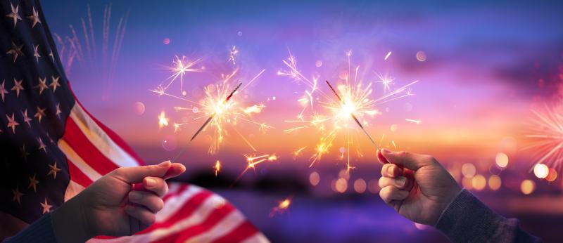 July 4 celebration hands holding sparklers with flag behind on left