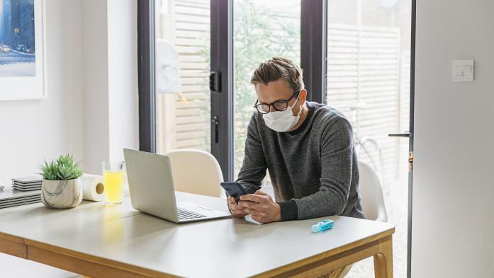 Man wearing mask looking at cell phone and laptop