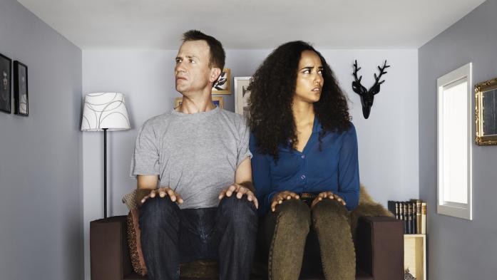 A confused couple sit in a tiny couch inside a tiny home