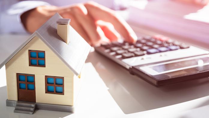 Photo of a persons hand using a calculator and a small house in the foreground