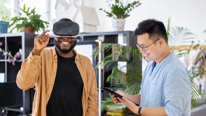 Colleagues testing out virtual reality device inside the metaverse - stock photo  African man wearing virtual reality glasses and gesturing with ale colleague standing by using digital tablet in office. Men testing out virtual reality device at office.