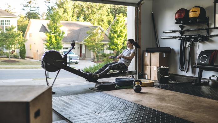 Photo of an active woman is exercising on a rowing machine in the home garage gym during covid-19 pandemic. Woman performing powerful pulls and focusing on endurance and cardio improvement.