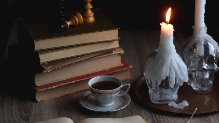 Stack of vintage books, cup of tea or coffee, lit candles, reading glasses and chess pieces on wooden table. Dark academia concept. Selective focus.