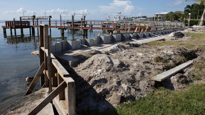 Seawall breakwall construction strengthening work on large body of water - stock photo  Seawall break wall concrete strengthening work construction site utilizing wood framing on large body of water with docks, boats and houses in the background on sunny.