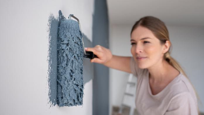 DIY woman painting a wall of her house blue using a paint roller - focus on foreground