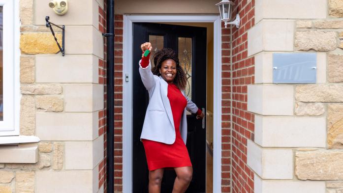 Three-quarter-length shot of a mature adult woman standing outside a vacant property. She is smiling happily at the camera, holding up a set of house keys in one hand whilst opening the door with the other. She is wearing smart casual clothing.