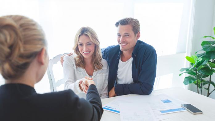 Real estate agent with couple shaking hands closing a deal. There is a digital tablet on the table. Couple are casually dressed. They are looking happy and smiling. Over the shoulder view inside a home.