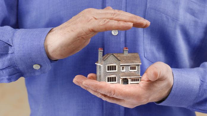 Model home in a man's one hand with his other hand covering it to suggest insurance
